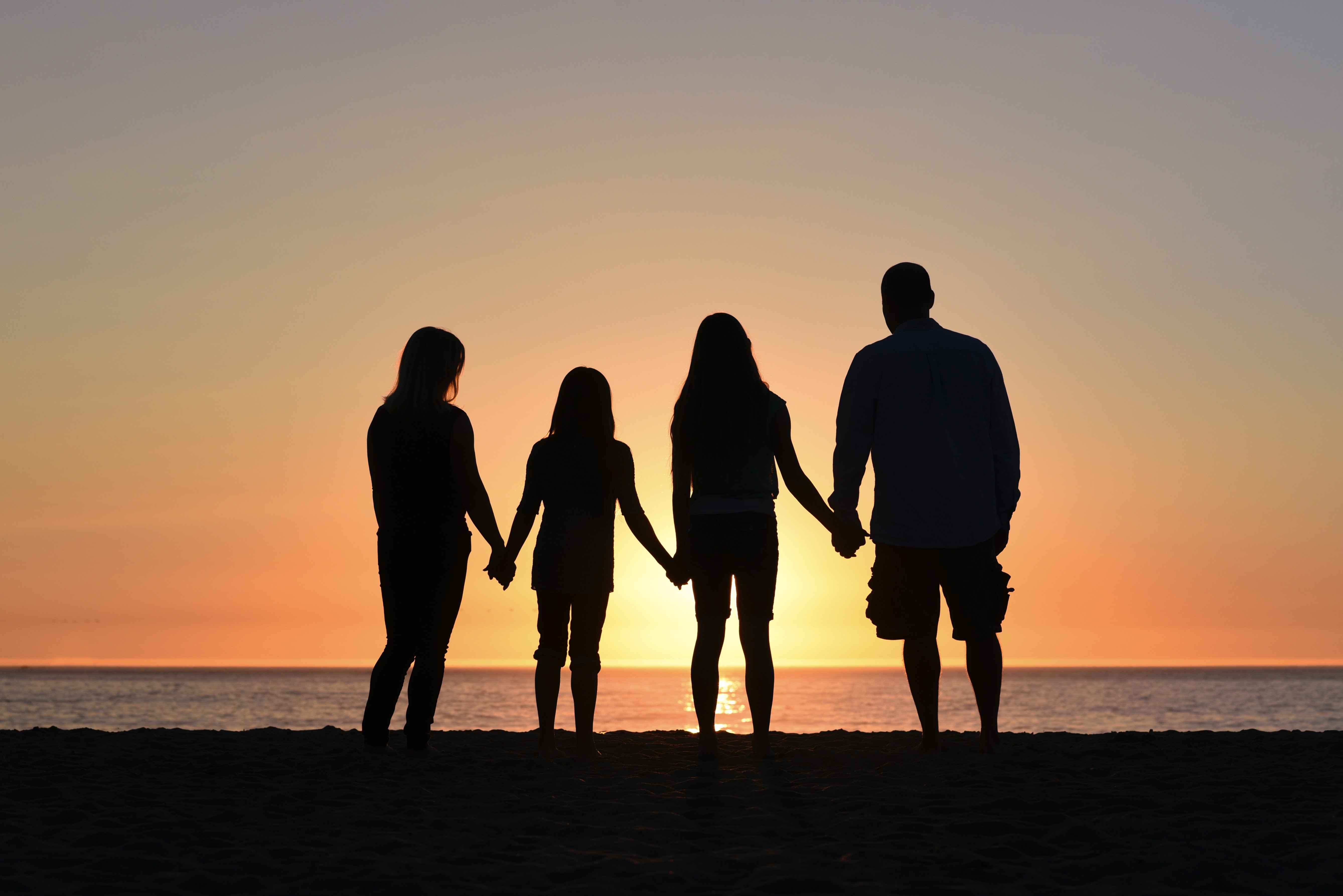 silhouette of mom dad and two kid in-between them holding hands while facing the sunrise some ocean water too